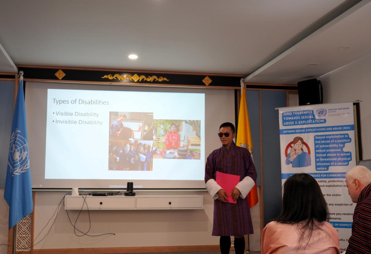 A UN Volunteer from FAO, delivering a presentation on disability inclusion, along with practical demonstrations on disability etiquette. He is a person with blindless, who is stood in front of the training participants delivering the presentation. 